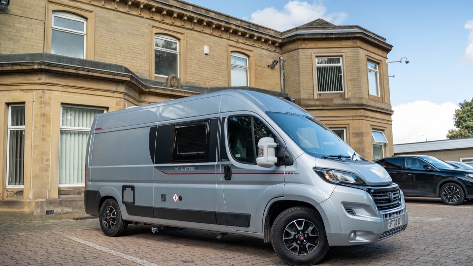A silver Fiat-based campervan parked on a block-paved courtyard outside Motorhome Pig's stone office building in Halifax