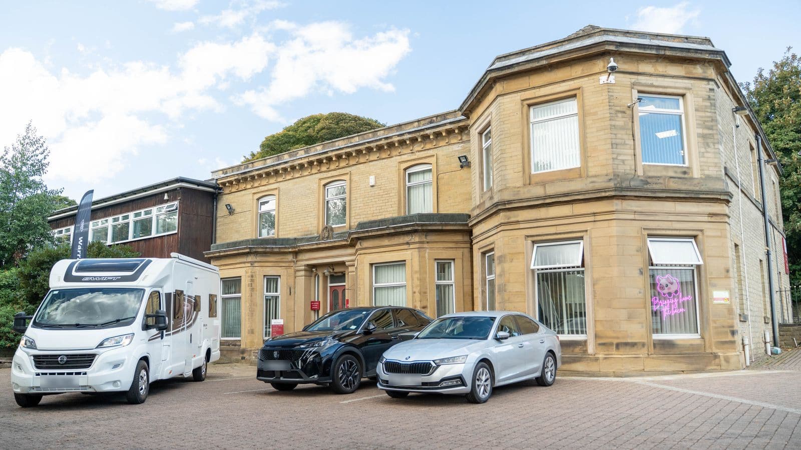 A Swift motorhome and two cars parked outside Motorhome Pig’s stone office building in Halifax, with the pink neon pig sign in the window