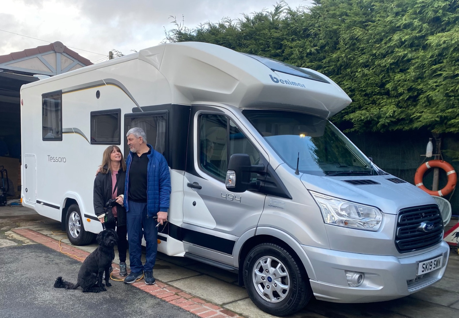 A couple and their dog beside the Benimar Tessoro motorhome they have just bought, on a driveway under an overcast sky
