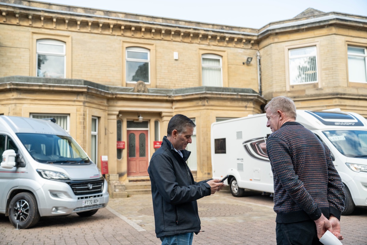 A man checking figures on his phone in the courtyard outside Motorhome Pig's office, with a campervan and a motorhome parked behind him