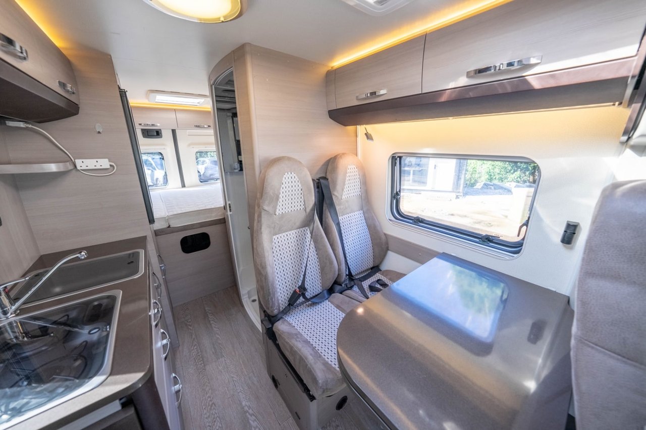 A bright, clean motorhome interior photographed in daylight, showing the kitchen, dinette and cab
