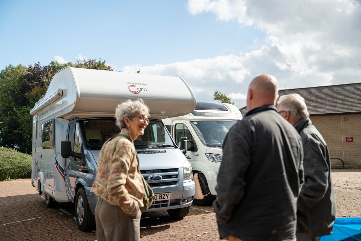 A smiling woman and two men talking beside a Ford-based Hymer motorhome in a sunny car park
