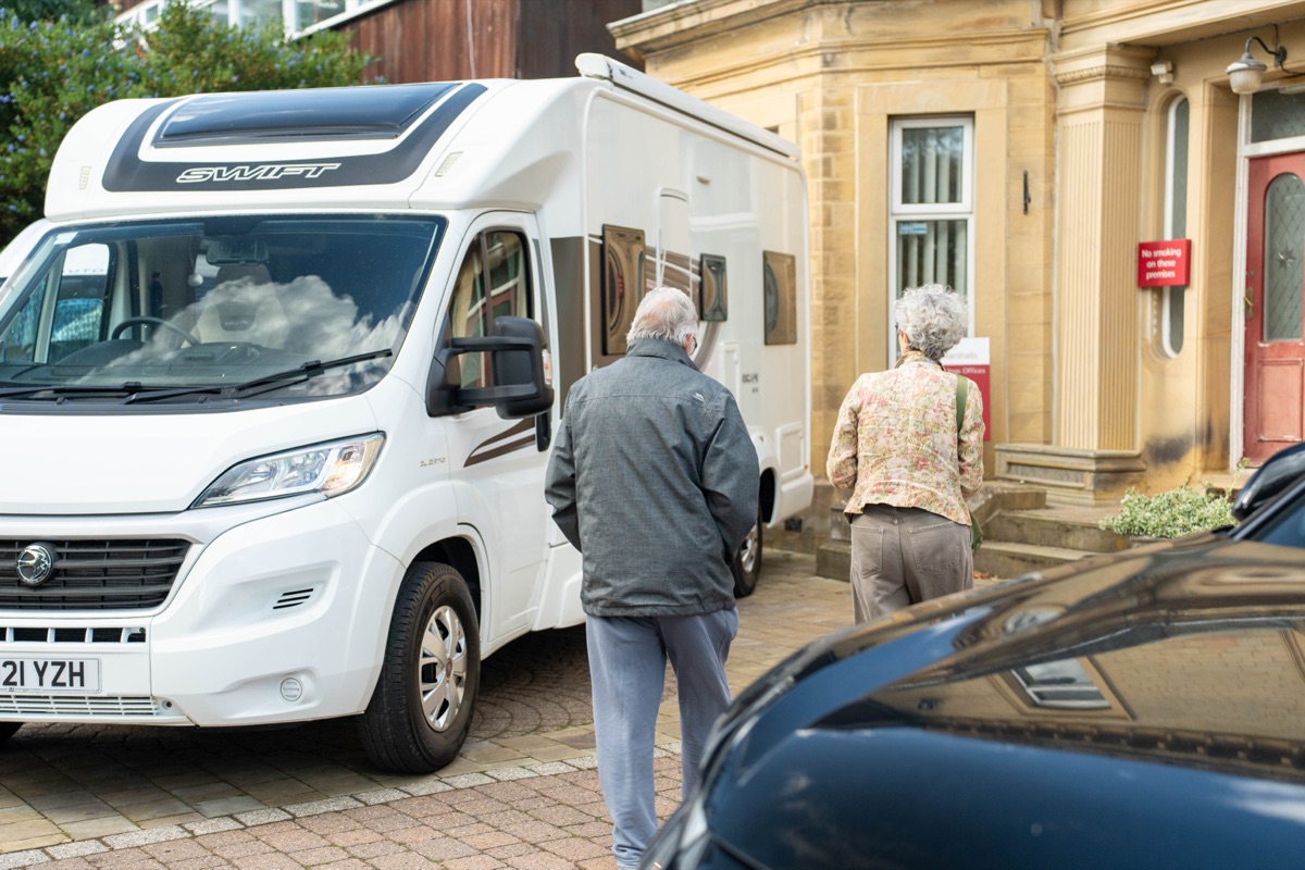 An older couple walking past a white Swift motorhome towards the entrance of a stone office building