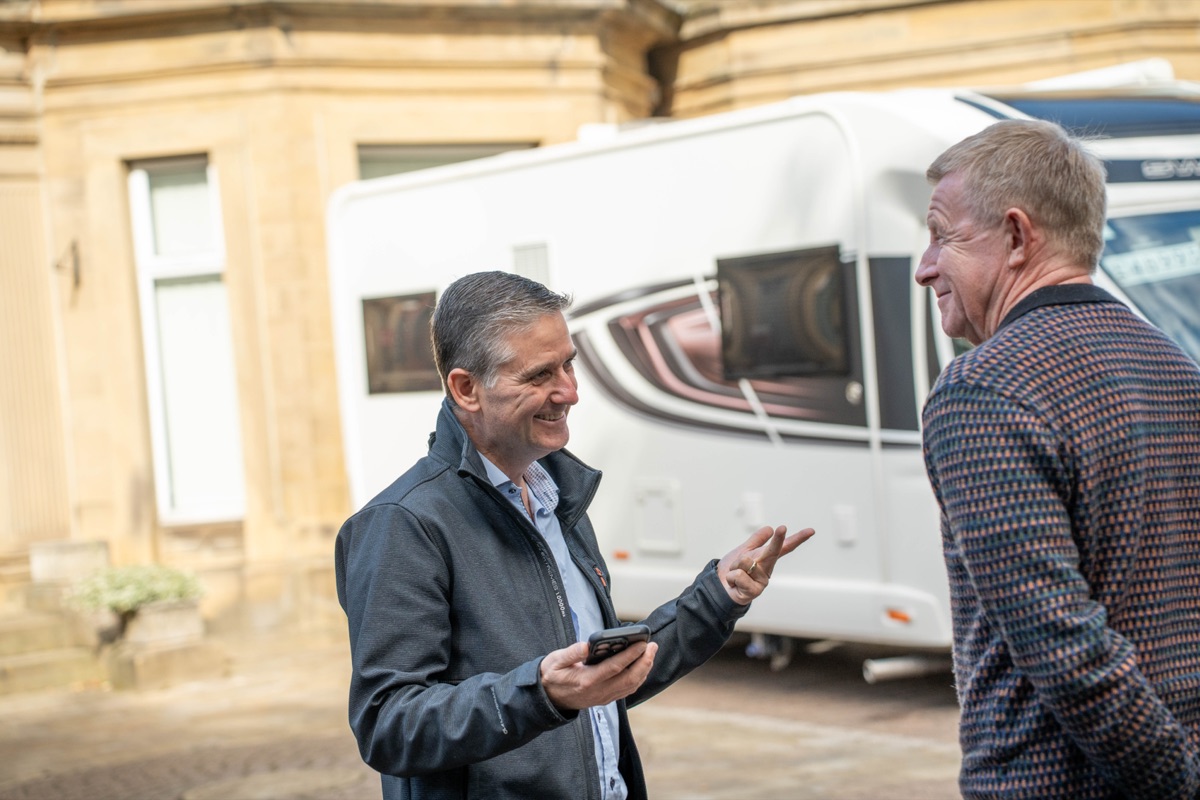 A member of the Motorhome Pig team chatting with a smiling man outside a stone building, with a white motorhome parked behind them