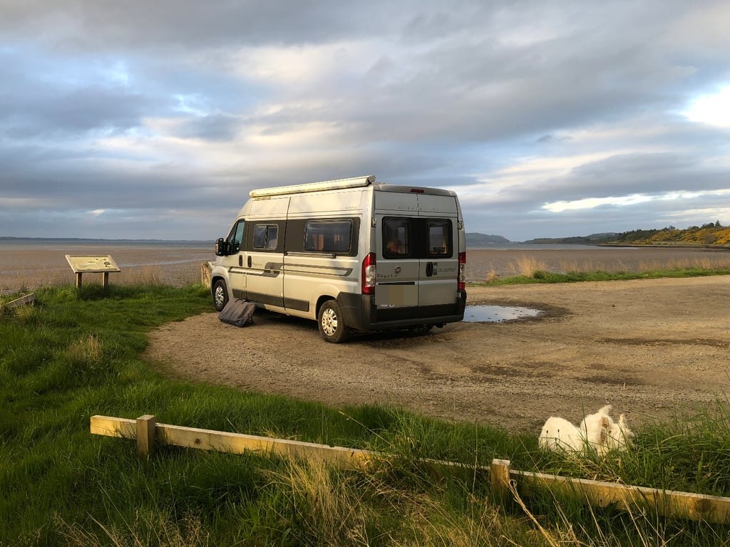The Autocruise Rhythm parked at a coastal viewpoint at dusk under a dramatic cloudy sky, with a small white dog in the grass nearby (number plate digitally obscured)