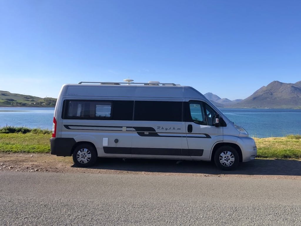Side profile of the silver Autocruise Rhythm campervan with 'Rhythm' badging, parked beside a Highland sea loch with mountains across the water
