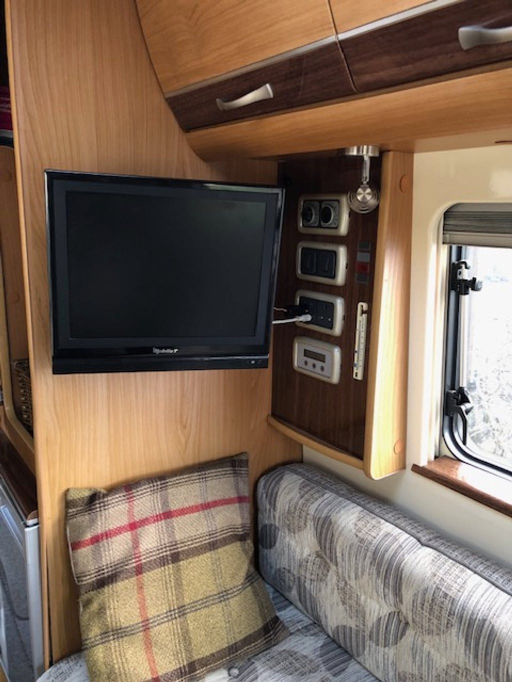 The interior rear lounge of the Autocruise Rhythm with upholstered seating, a tartan cushion, wood cabinetry and a wall-mounted TV