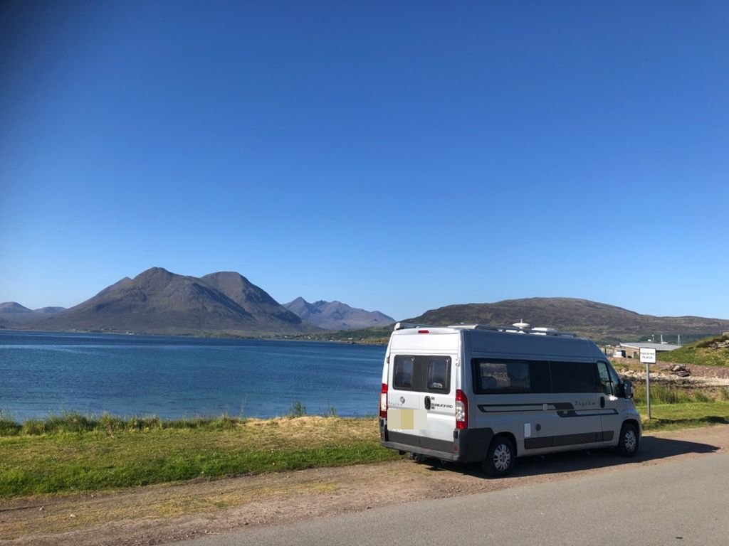 The Autocruise Rhythm parked at a loch-side viewpoint with the Cuillin mountains of Skye in the distance under a clear blue sky (number plate digitally obscured)