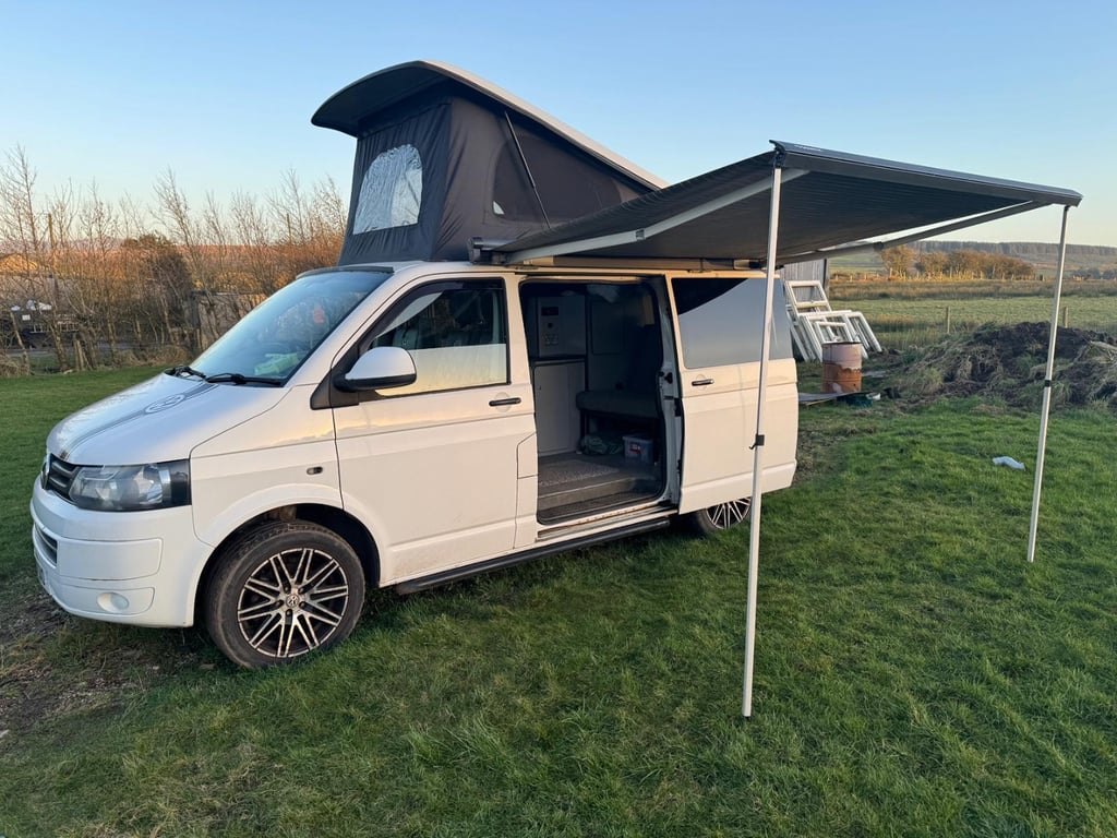 Side view of the white VW Transporter campervan pitched on grass at dusk with its pop-top roof raised, side door slid open showing the galley, and the wind-out awning extended over the grass