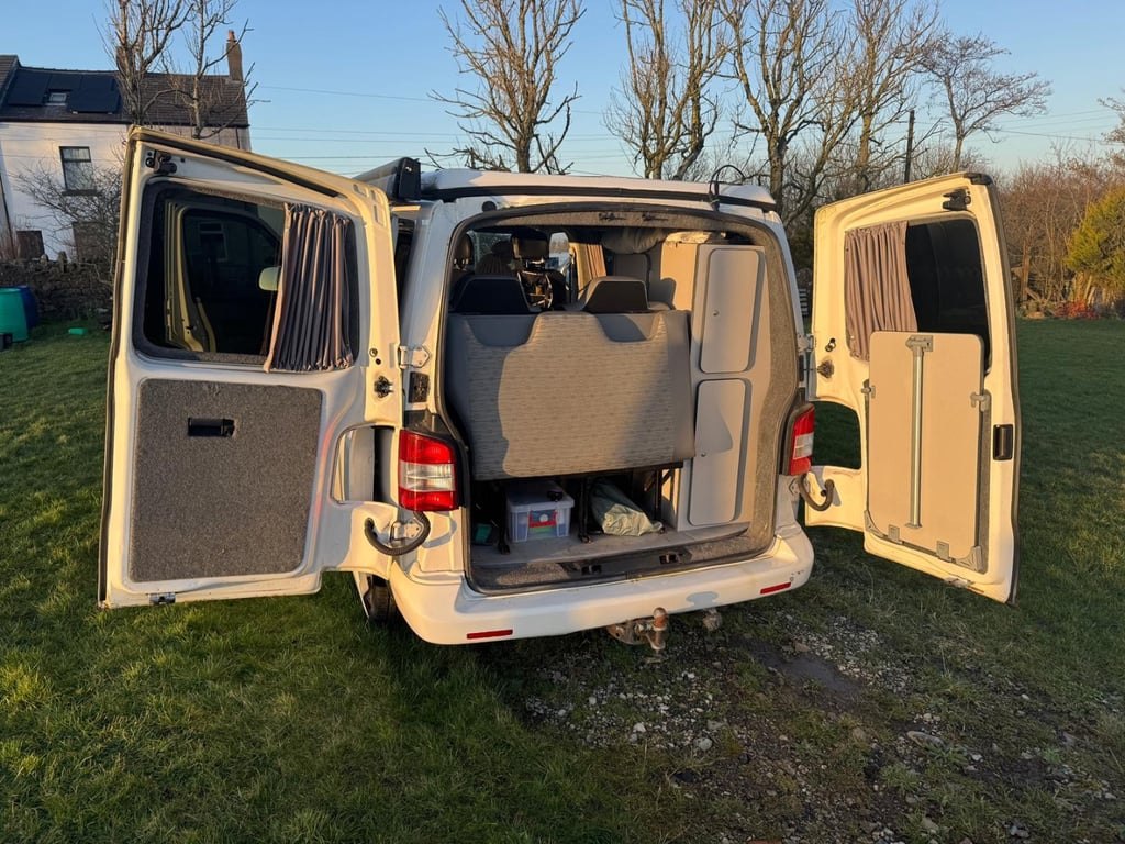 Rear view of the VW Transporter campervan on grass with both rear barn doors open, showing the rock-and-roll bed seat back, curtains and interior panelling