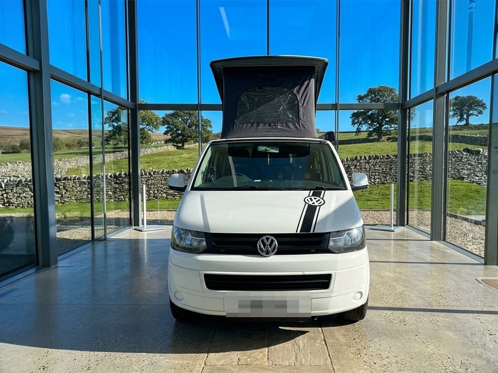 A white 2011 Volkswagen Transporter T5.1 campervan with its pop-top roof raised, photographed head-on inside a glass-walled building with Cumbrian fields and a dry stone wall behind (number plate digitally obscured)