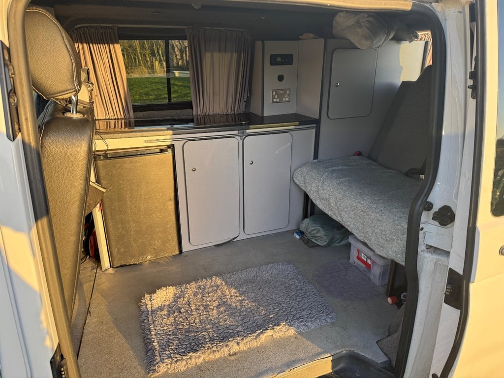 Interior of the VW Transporter campervan through the open side door, showing a compact galley with worktop, cupboards, a fridge and the rock-and-roll bench seat behind