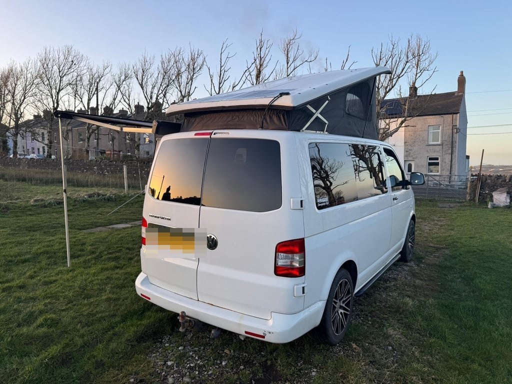 Rear view of the white VW Transporter campervan parked on grass with its pop-top roof raised and awning partially extended, houses and a hillside behind (number plate digitally obscured)