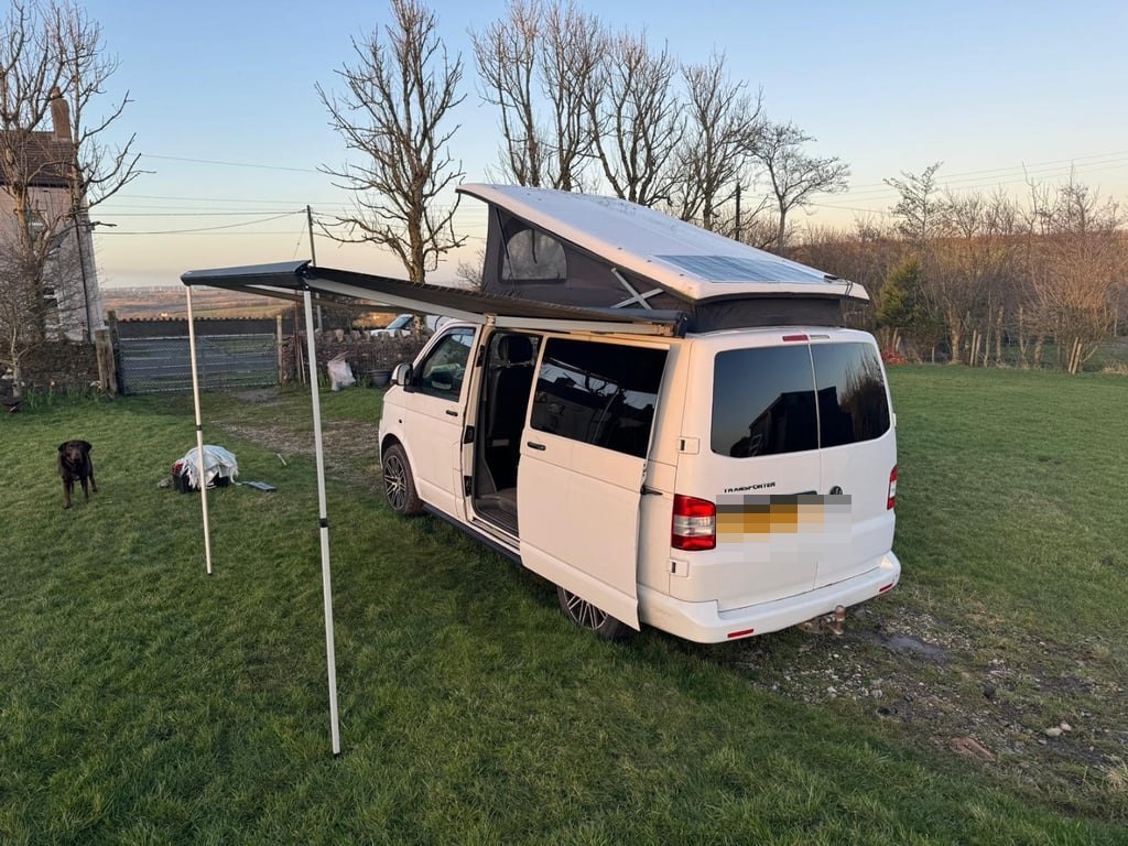 Rear three-quarter view of the white VW Transporter campervan on grass at sunset, pop-top roof raised with a solar panel on it, wind-out awning extended, and a dog in the background (number plate digitally obscured)