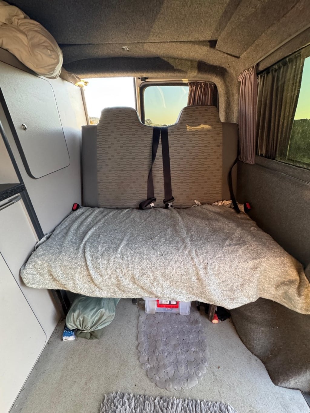 The rock-and-roll bed in the VW Transporter campervan folded flat into a double bed with a grey mattress topper, seat belts visible on the backrest