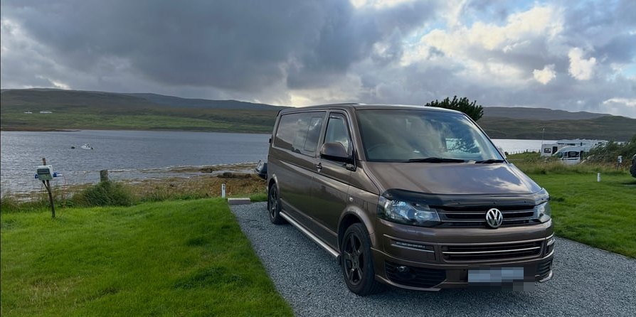 A brown Volkswagen Transporter campervan pitched on a gravel hardstanding beside a sea loch, with hills and low cloud behind
