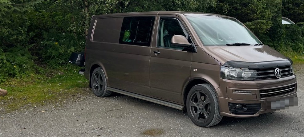 The full side profile of a brown long-wheelbase Volkswagen Transporter campervan parked on a gravel track in front of dense green trees