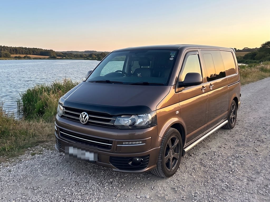 A brown Volkswagen Transporter campervan parked on a gravel track beside a lake at sunset, with reeds in the foreground and the sky glowing orange behind
