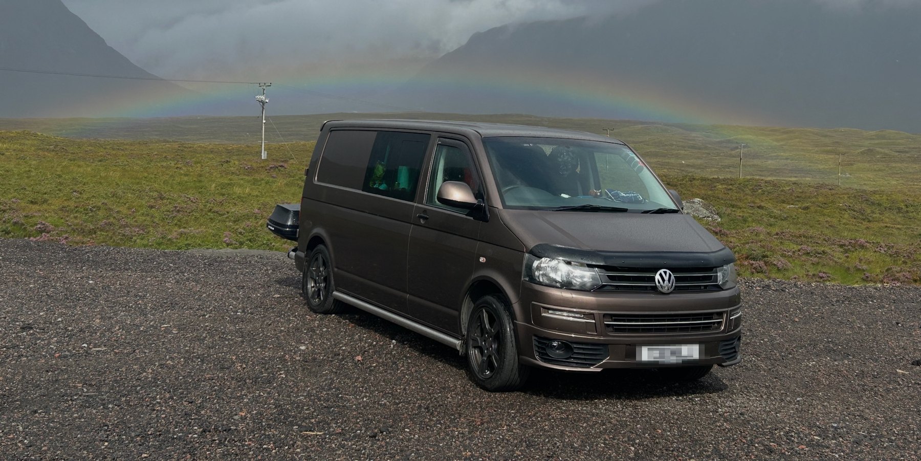 A brown long-wheelbase Volkswagen Transporter campervan parked on a gravel layby in the Scottish Highlands, with a rainbow arcing over moorland and a dark mountain behind