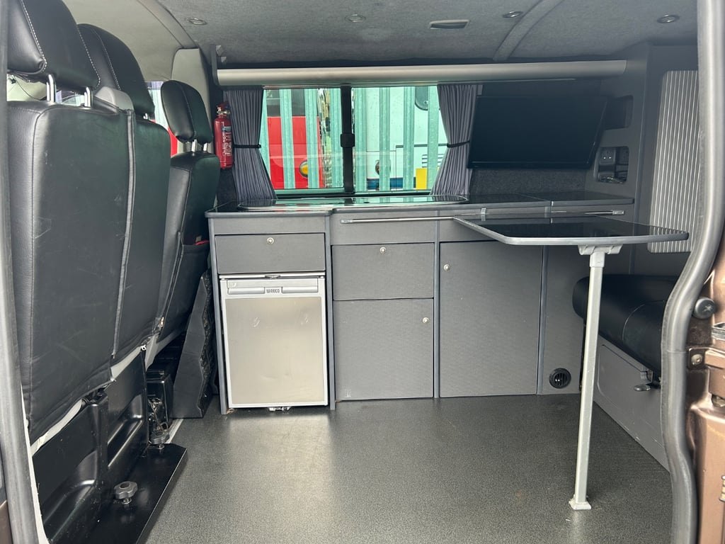 The campervan's kitchen unit photographed from the sliding door, showing the worktop, the folding table stowed and the striped window blinds