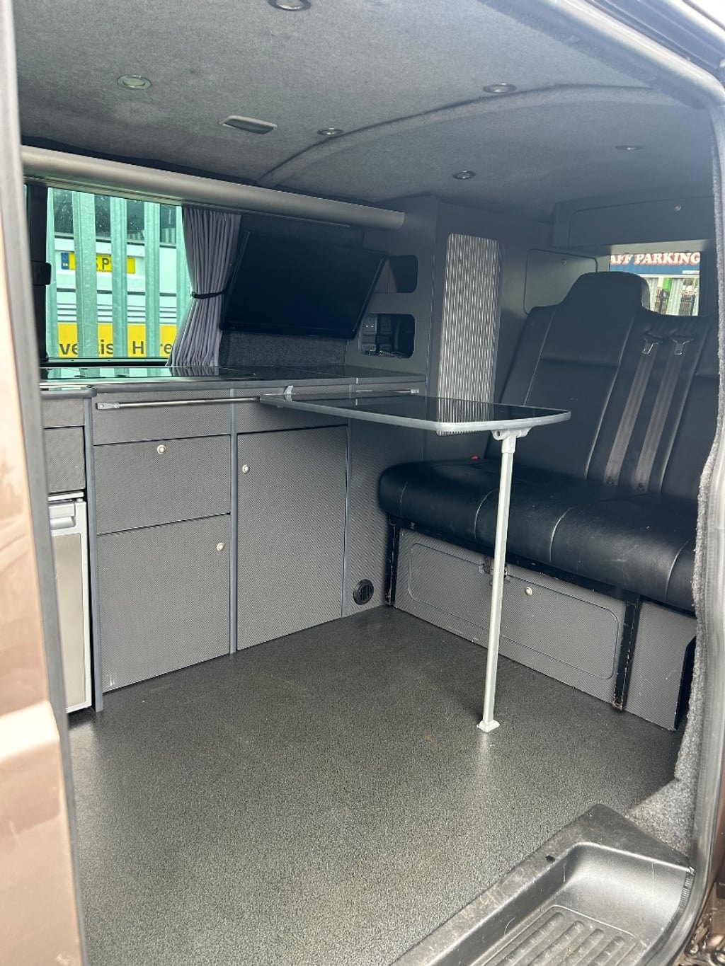 The interior of the campervan looking rearward, showing a grey kitchen unit with a worktop, a folding table and a bench seat in dark leather