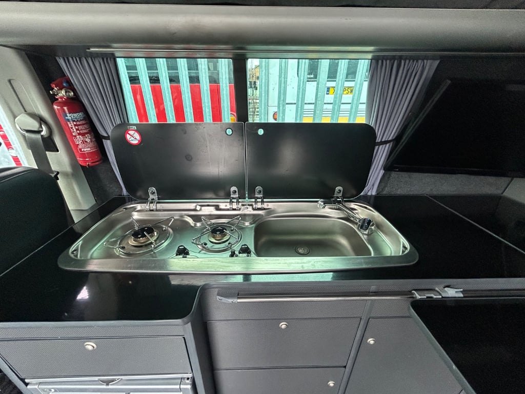 A stainless steel sink and gas hob set into the campervan's kitchen worktop, with a striped blind at the window above