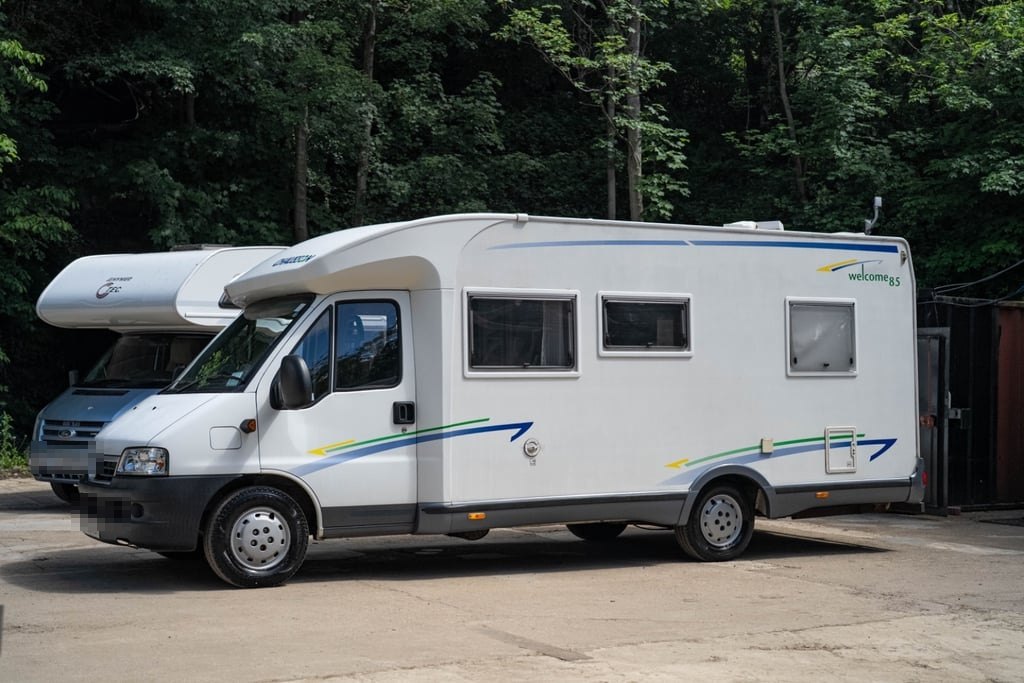 A white 2006 Chausson Welcome 85 coachbuilt motorhome parked on a hardstanding in front of woodland at Motorhome Pig's Halifax site (number plates digitally obscured)