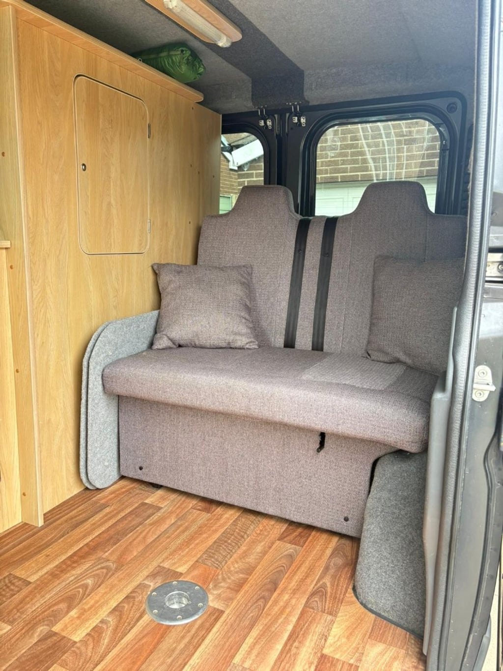 The grey bench seat inside the campervan in its upright position with cushions, next to wooden cabinets and a wooden floor