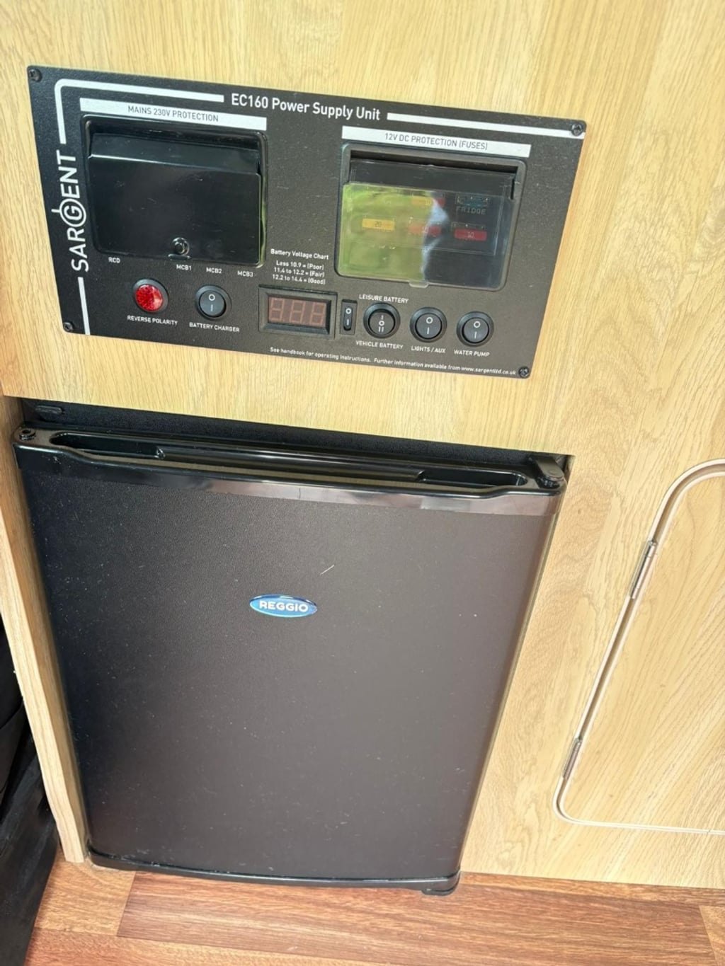 The campervan's black fridge set into wooden units, with a Sargent electrical control panel mounted above it