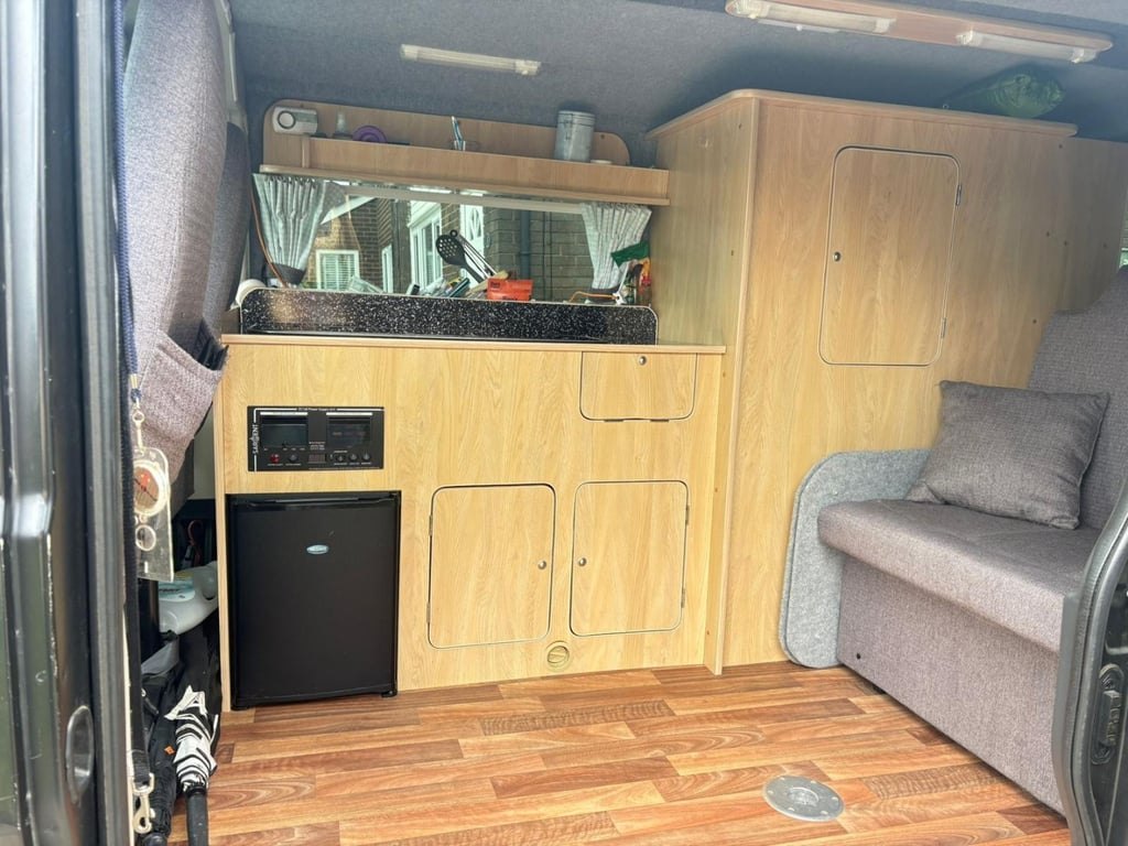The interior of the Peugeot Boxer campervan seen through the open sliding side door, showing the wooden kitchen units, black fridge, control panel and grey bench seat
