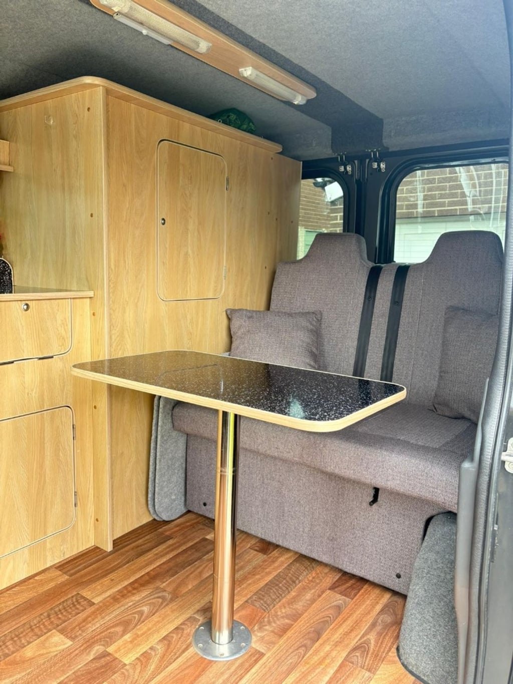 The campervan interior with a black pedestal table set up in front of the grey bench seat, which has visible seat belts