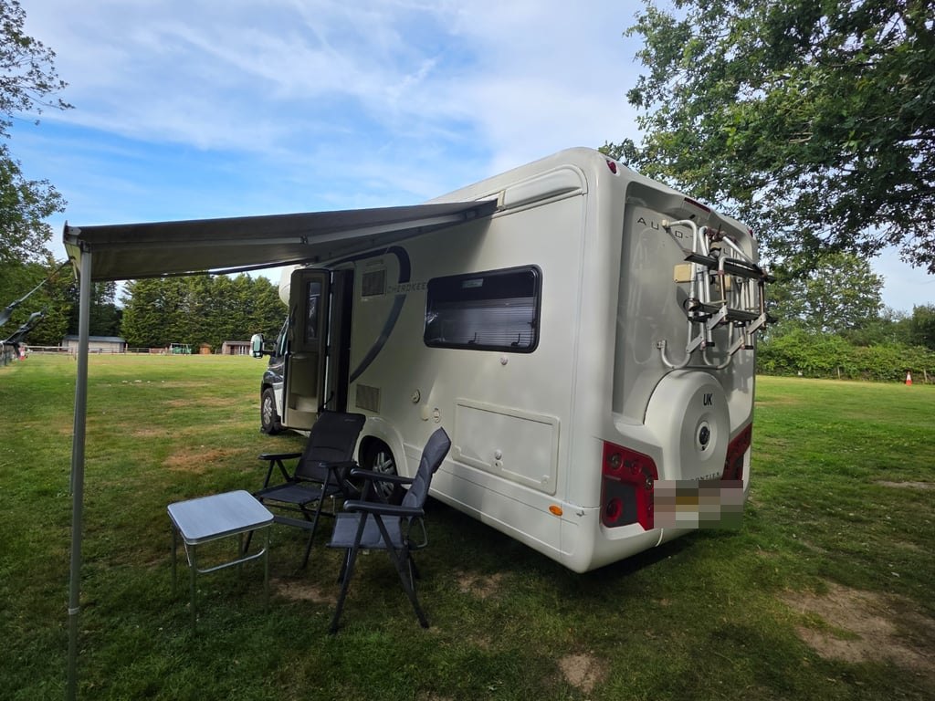Rear three-quarter view of the Auto-Trail Cherokee on a grass pitch with its wind-out awning extended over a table and two camping chairs (number plate digitally obscured)