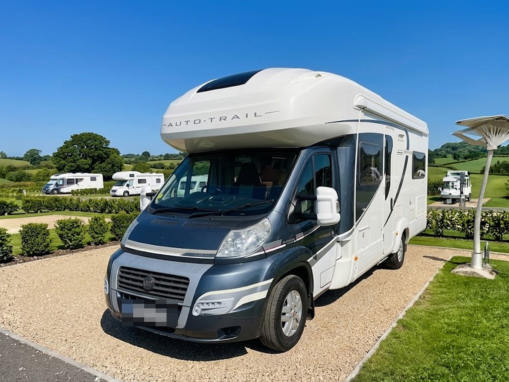 A 2013 Auto-Trail Cherokee motorhome with a grey Fiat Ducato cab and white coachbuilt body, parked on a gravel campsite pitch under a clear blue sky with green hills behind (number plate digitally obscured)