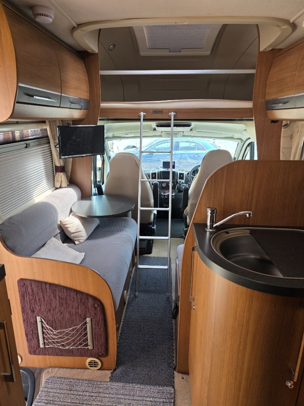 Interior of the Auto-Trail Cherokee looking forward from the rear, showing the side settee, the round table, a wall-mounted TV, the corner kitchen with a round stainless sink, and the cab beyond