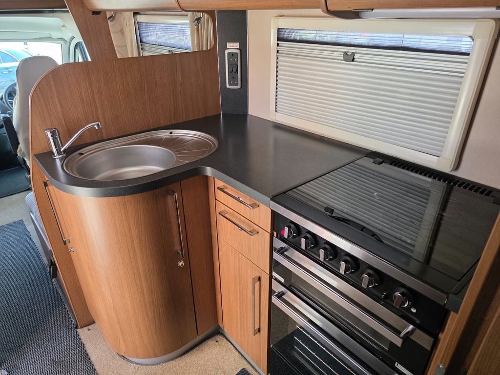 The Auto-Trail Cherokee galley with a round stainless sink in a dark worktop, a four-burner gas hob with a glass lid and an oven and grill below, in light wood cabinetry