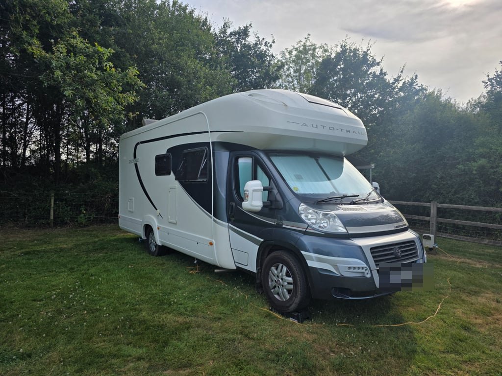 Front three-quarter view of the Auto-Trail Cherokee on a grass pitch at dusk, hooked up to an electric bollard by a yellow cable, with trees behind (number plate digitally obscured)