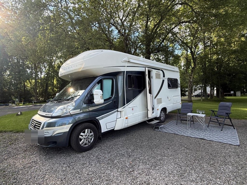 The Auto-Trail Cherokee pitched on a gravel campsite in the evening sun, with the habitation door open, a mat, a folding table and two camping chairs set out alongside (number plate digitally obscured)
