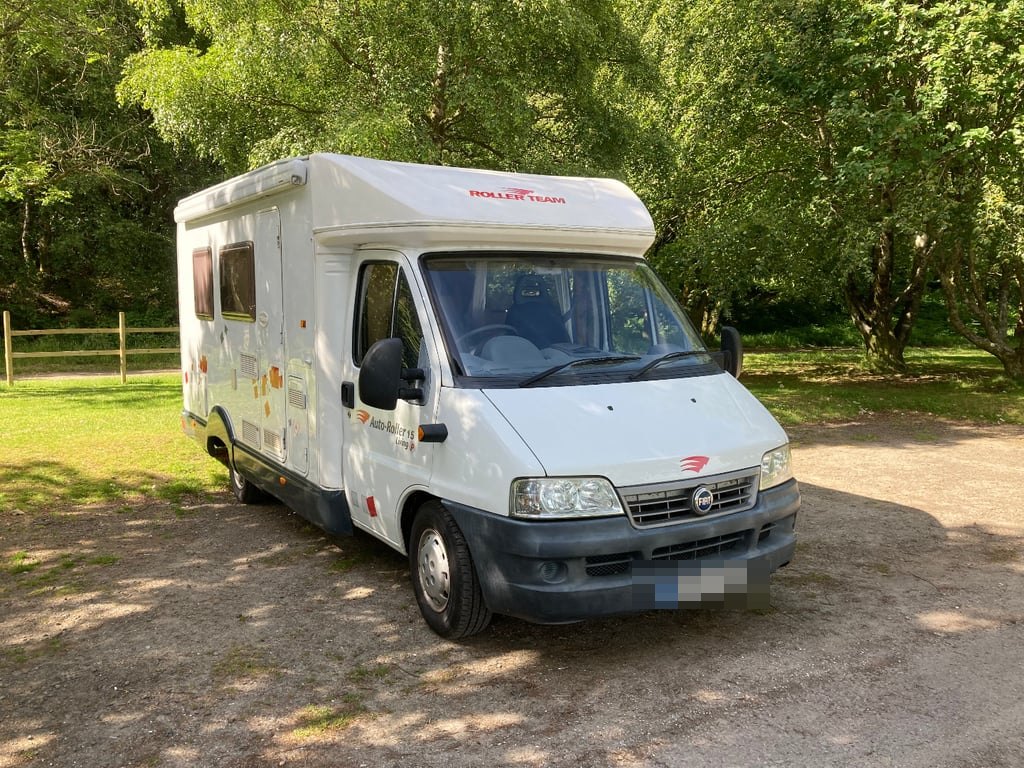 The Roller Team Auto-Roller 15 photographed from the front three-quarter angle in a wooded car park, showing the Fiat Ducato cab and the over-cab section (number plate digitally obscured)