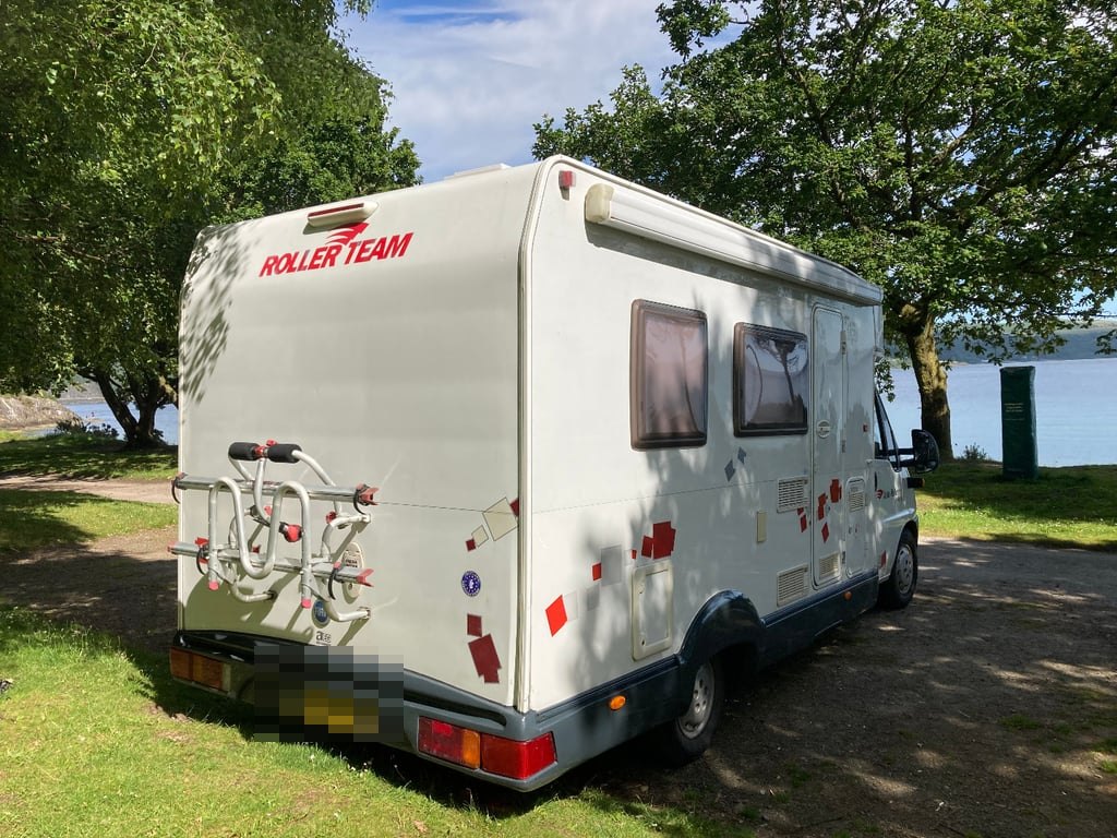 The rear three-quarter view of the Roller Team Auto-Roller 15 parked on grass beside a loch, showing the bike rack mounted on the rear panel (number plate digitally obscured)