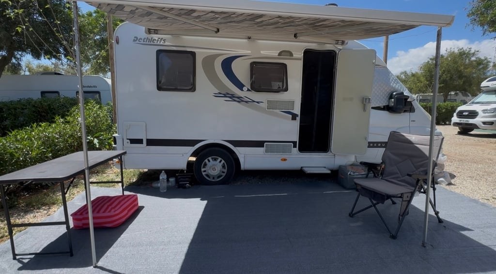 A white 2007 Dethleffs Globebus motorhome pitched on a sunny campsite with its wind-out awning extended, a table and camping chairs set up alongside (number plate digitally obscured)