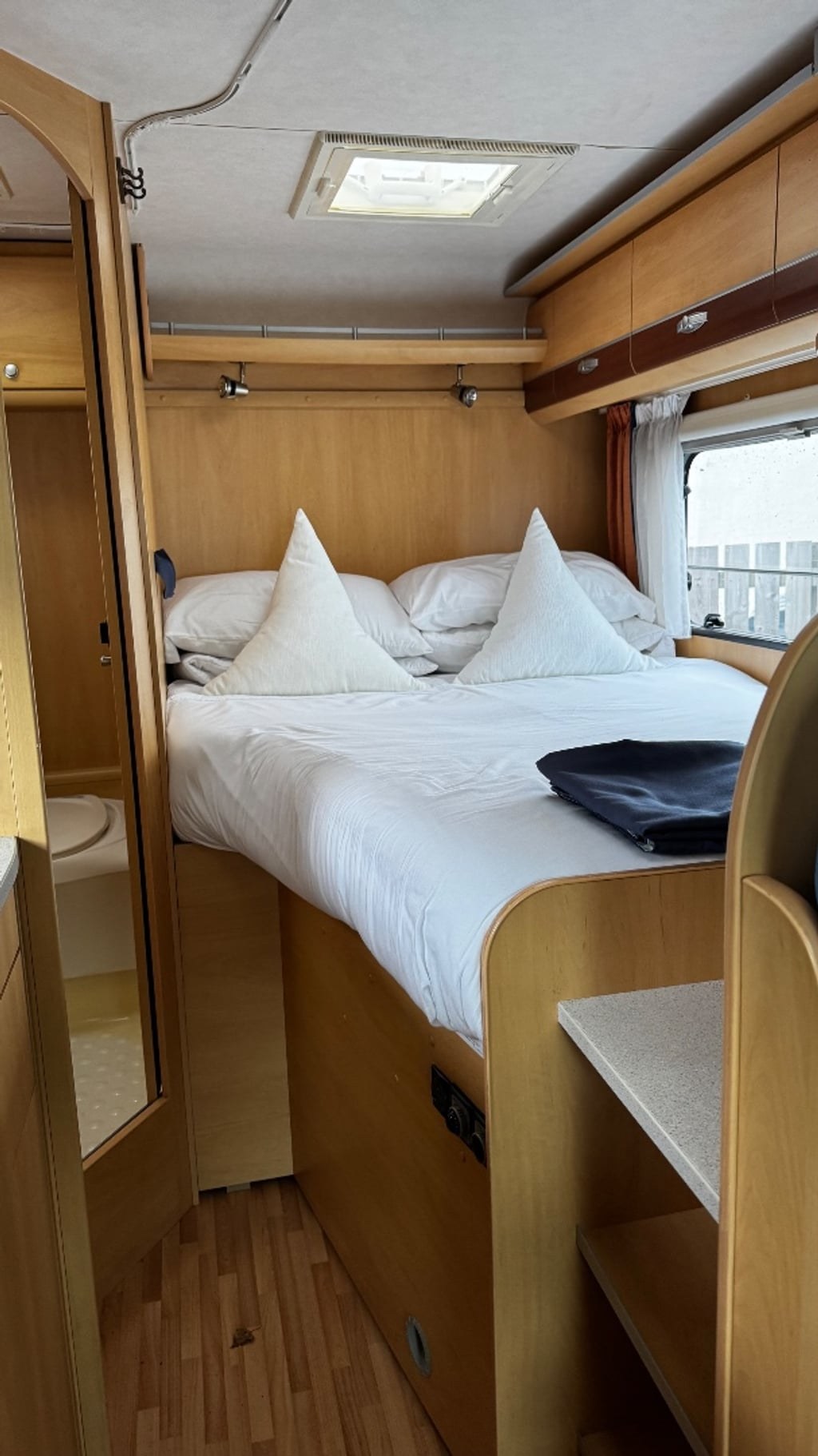 The fixed rear double bed of the Dethleffs Globebus, made up with white bedding and pillows under an overhead locker and rooflight