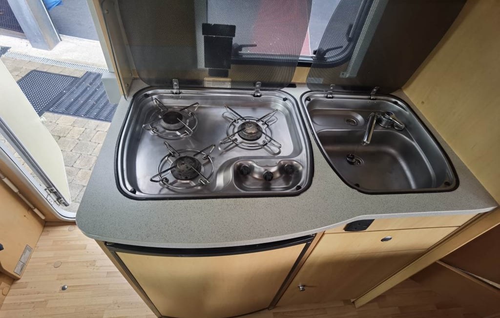 The Dethleffs Globebus kitchen with a three-ring stainless gas hob and a separate stainless sink set into a grey worktop, with light wood cabinetry