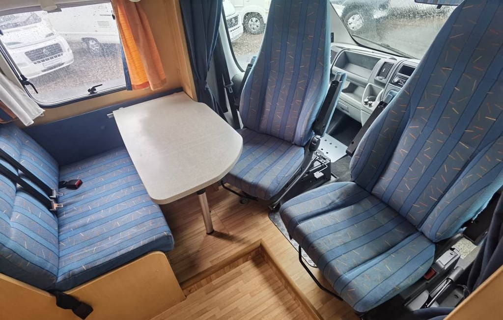The interior of the Dethleffs Globebus looking forward, with the swivelled cab seats and dinette in blue upholstery and a light wood-finished interior