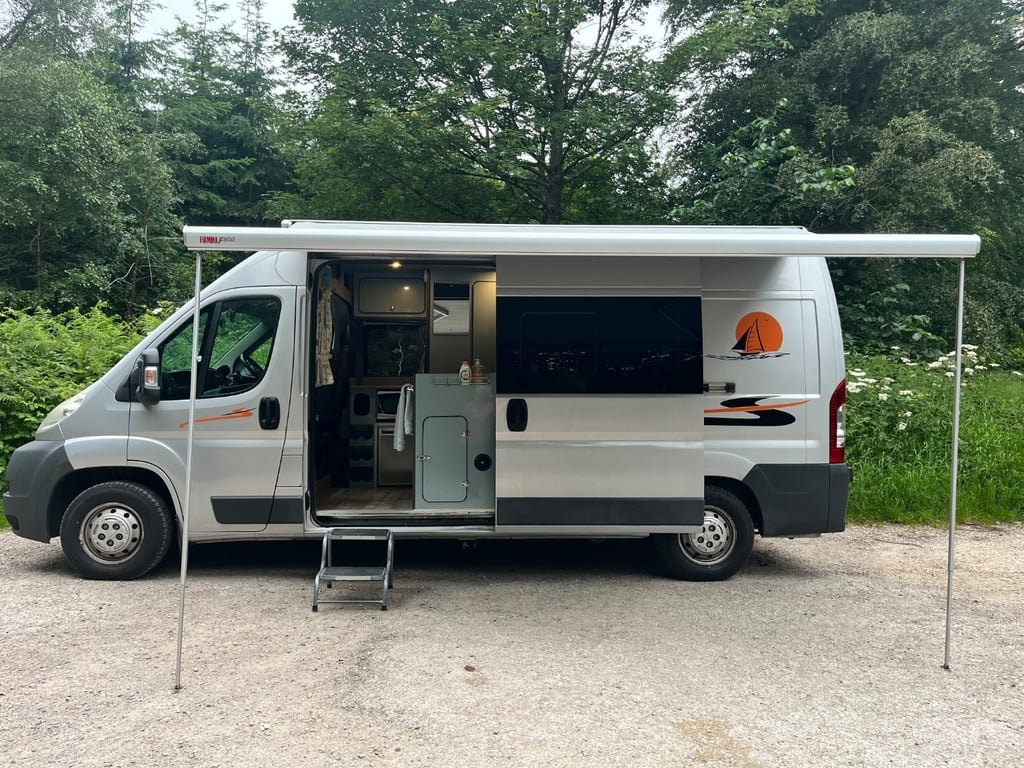 The silver campervan parked on a woodland track with its wind-out awning extended over the open side door