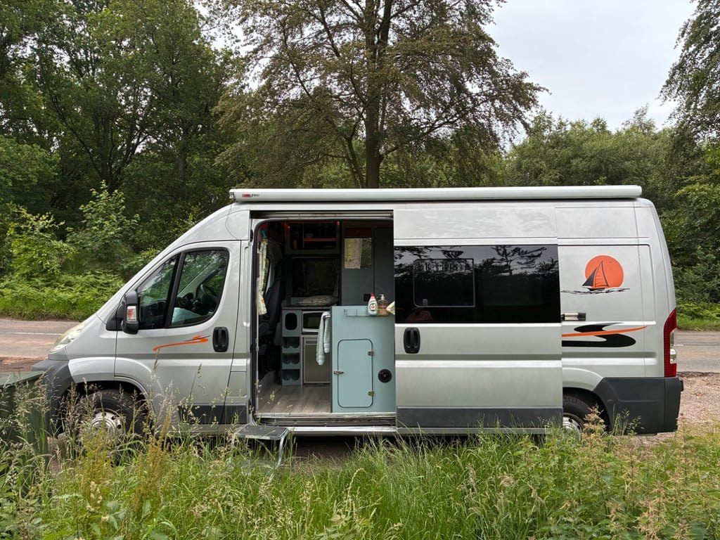 The self-converted campervan parked on a woodland track with the side door slid open, showing the galley and wet-room door inside