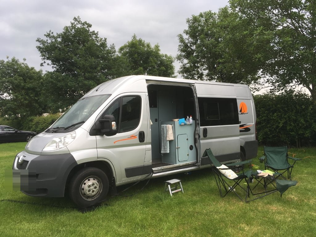 The self-converted campervan pitched on grass with camping chairs and a table set out beside its open side door
