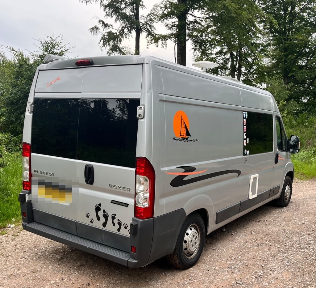The rear three-quarter view of the campervan on a woodland track, showing a sailing-boat graphic and the name Connie on the tailgate, with the number plate digitally obscured
