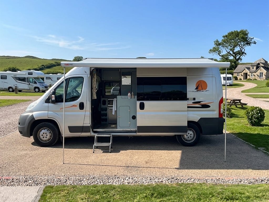 A silver self-converted Peugeot Boxer campervan pitched on a sunny campsite with its wind-out awning extended, side door open and step down