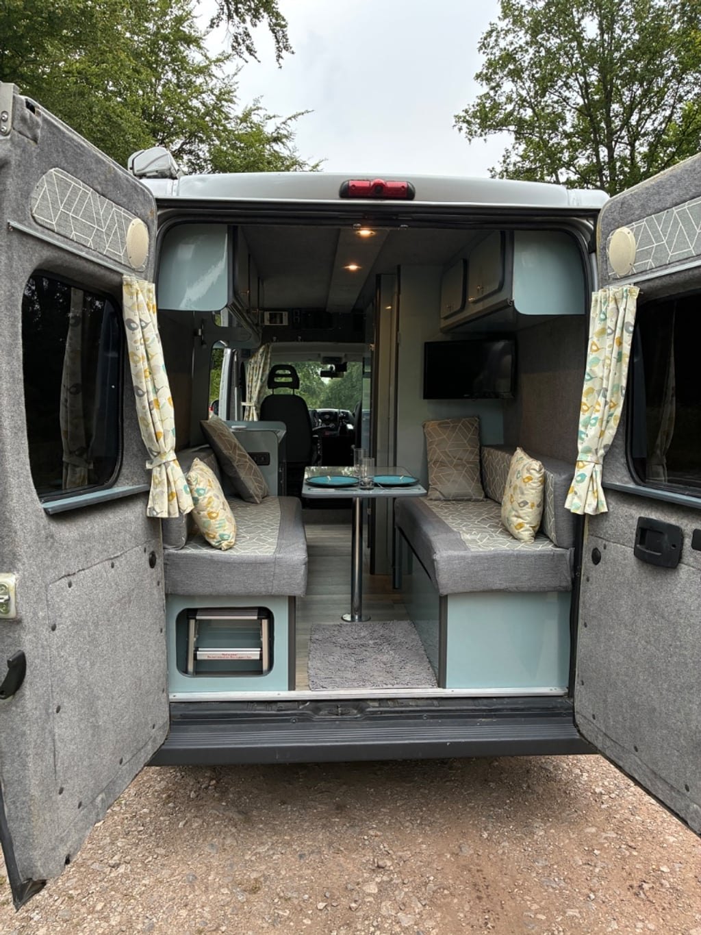 The rear doors of the campervan open to show a facing pair of upholstered seats, a pedestal table set for two, and overhead lockers