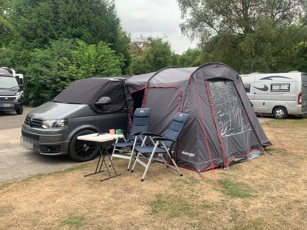 The campervan pitched on a campsite with a large grey Vango driveaway awning attached, camping chairs and a table set out on the grass (number plates digitally obscured)